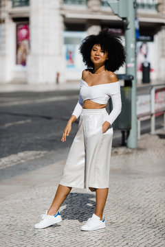 Beautiful African American woman in white fashionable outfit standing with hands in pockets on roadside against urban background