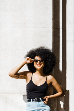 Young ethnic woman in jeans and tank top leaning on wall and smiling at camera outdoors