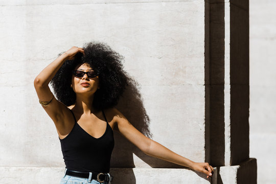 Young ethnic woman in jeans and tank top leaning on wall and smiling at camera outdoors