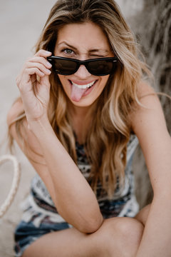 Close-up portrait of pretty blonde woman in black sunglasses sitting on sand and looking at camera sticking out tongue and winking