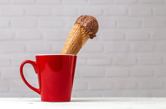 Delicious waffle cone with brown ice cream scoop served in mug on white table against brick wall