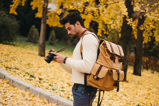 Side view of handsome young photographer standing in autumn park and watching pictures on camera