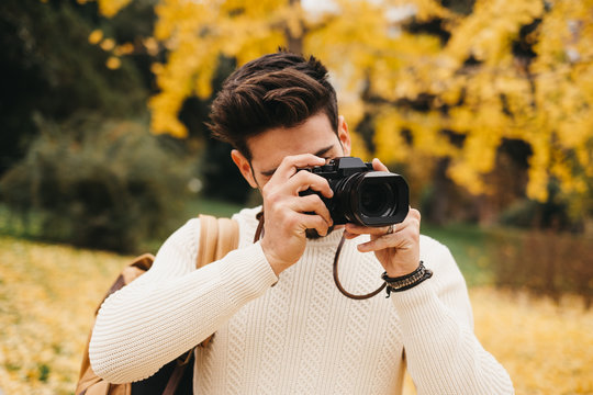 handsome young photographer standing in autumn park and taking pictures with camera
