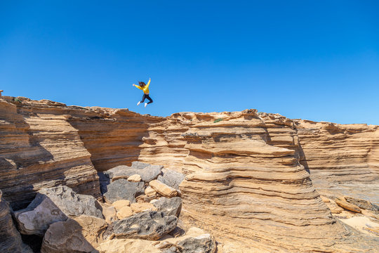 Young woman in yellow sweatshirt jumping with spread hands over deserted sandstone rocks on sunny day