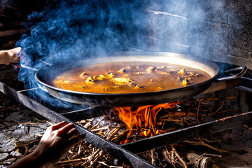 Crop unrecognizable hands holding big iron pan with boiling broth for cooking paella over open fire with wood
