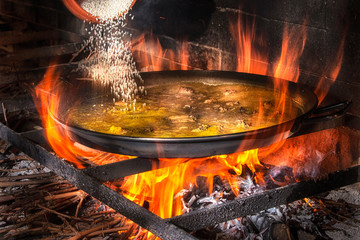 Adding rice into big iron pan with boiling broth for cooking paella over open fire with wood
