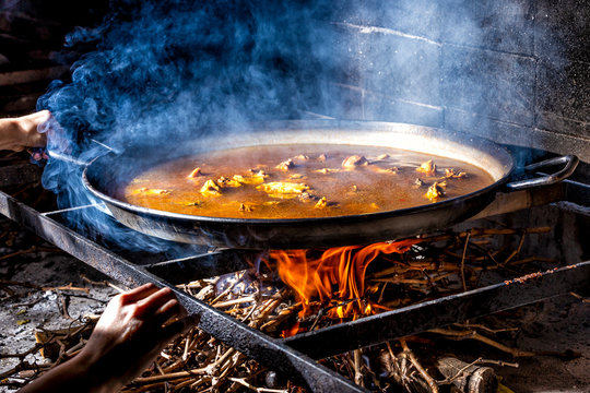 Crop unrecognizable hands holding big iron pan with boiling broth for cooking paella over open fire with wood