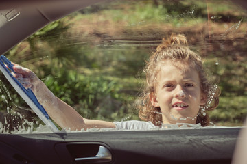 Smiling kid having fun with foam washing front glass of car by hand