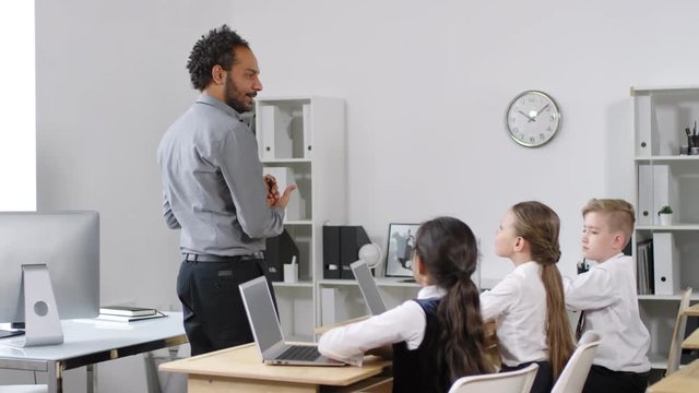 Medium shot of vivacious young African American male teacher pacing around in front of attentive preteen schoolchildren in uniform, sitting at desks with laptops on them, and explaining new material