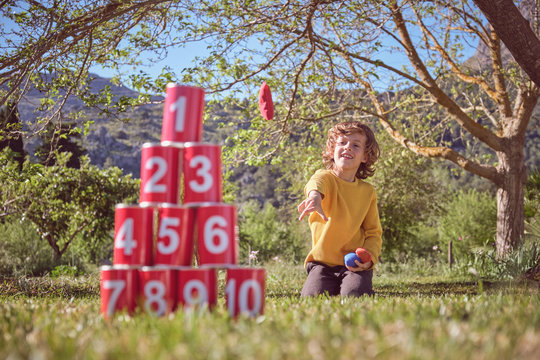 Cheerful kid kneeling and throwing balls in pyramid of red tin cans on nature background