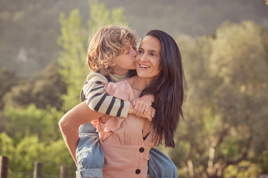 Laughing mom and son having fun doing piggyback on countryside and woman keeping son on back