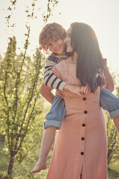 Laughing mom and son having fun doing piggyback on countryside and woman keeping son on back
