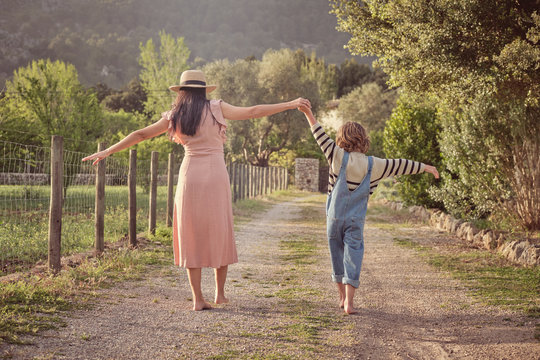 Back view of mother and son holding by hands and walking barefoot along rural road in bright day