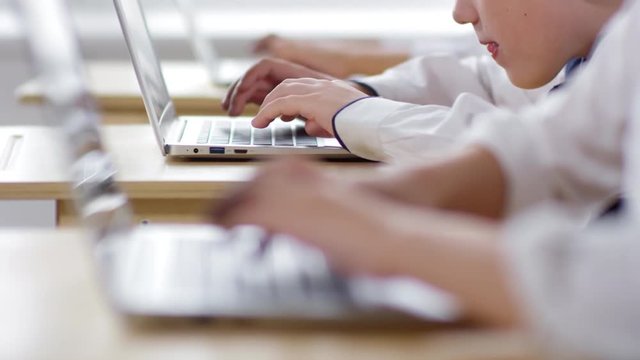 Close-up rack focus hands shot of schoolchildren, wearing uniform white shirts, sitting in classroom during practical computer education lesson and typing on laptops