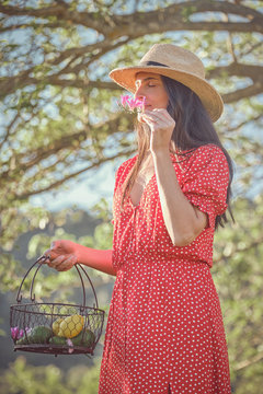 Barefoot young attractive woman in hat and red dress walking in green garden with basket of fruits and holding a fresh flower in summer day