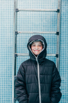 Smiling male kid in warm jacket standing on background of pool wall and looking at camera