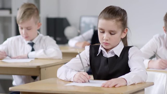 Waist-up shot of 9-year-old Caucasian schoolgirl, wearing uniform white shirt and pinafore, sitting at desk in classroom and filling out answer sheet during module test, and classmates in background