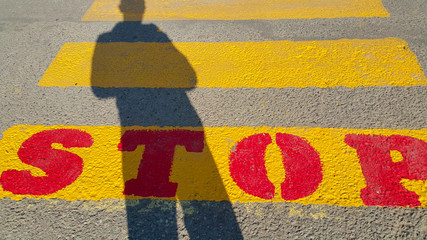 a person stands at the beginning of a pedestrian crossing, where it is written stop and waits for the passage time, on the yellow lines drawn on the asphalt, the shadow of a person is clearly visible,