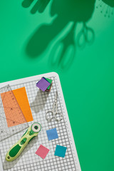 Rotary cutter, ruler, bright square pieces of fabric, stack of bright fabric square pieces, scissors on craft mat, green background