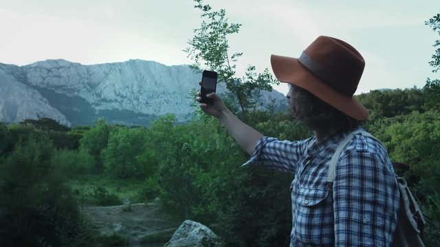 A young guy hiking in the woods on a background of mountains. Makes a photo of the mountains on a smartphone. Active lifestyle. Wearing a checkered shirt and a traveler's hat. Hiking travel concept.