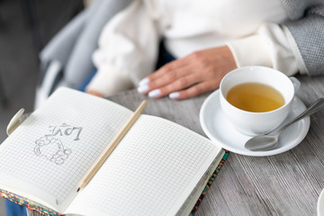 drawing of heart and word love are drawn in pencil in notebook, which lies on table in cafe next to cup of tea