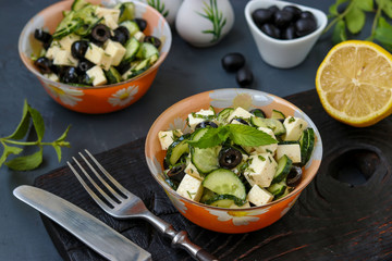 Healthy salad with cucumbers, feta and olives, with olive oil and greens, located in bowls against a dark background, horizontal photo