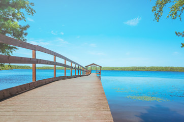 Pier on the lake with calm water under a bright blue sky. Soothing calm minimalistic landscape. Trees surround the pond. Place for text.