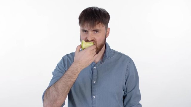 Medium shot of young bearded man standing isolated on white background, eating green apple and looking at camera