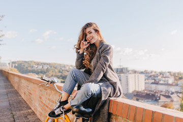 Well-dressed lady in good mood playfully posing on city background in warm weather. Charming dark-haired girl in coat sitting on the street under blue sky.