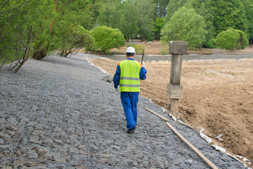 a builder in a blue uniform and a white helmet, carries a wooden beam on a stone embankment, the rear view, there is space for advertising