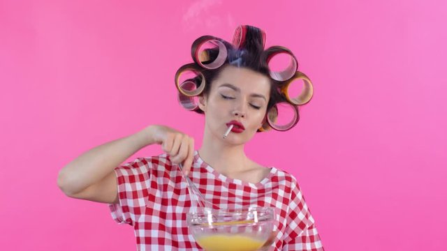 Medium shot of young woman wearing hair rollers, smoking cigarette and whisking egg yolks on pink background