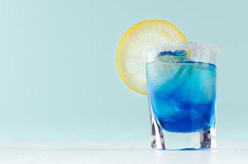 Exotic cold shot glass cocktail with yellow lemon slice, blue curacao, ice cubes, salt rim on soft light mint color background and white wood board.