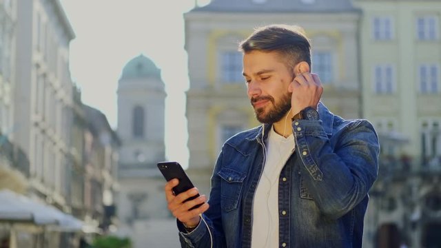 Cheerful good-looking bearded man in trendy jacket listening inflammatory music on headphone and dancing on the street background