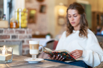 Young Caucasian calm girl sits in cafe and reads book. Latte on table. Quiet calm morning atmosphere. Comfort