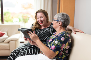 Two women sitting on sofa watching social media and funny viral videos on a tablet. The overweight woman is laughing and the  elderly woman with short gray hair is holding a tablet