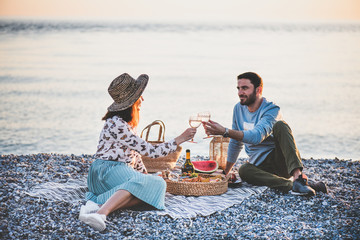 Summer beach picnic at sunset. Young happy couple having weekend picnic outdoors at seaside with bottle of sparkling wine, fruit and tray of tasty appetizers, drinking wine and looking to each other