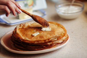 Stack of hot classic Russian thin pancakes. Rustic style. Traditional for the Russian pancake week - Maslenitsa. Female hand puts the butter on the hot pancakes.