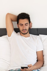Young handsome man sitting on the bed. He uses a mobile phone. The problem of dependence on mobile social networks.