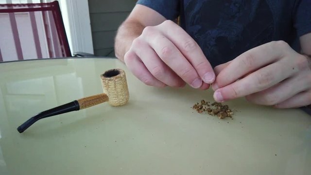 Closeup of man's hands tearing marijuana plant to pack in a weed smoke corncob pipe bowl.