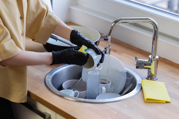 A young woman cleans up in the kitchen, washing dishes. She is tired and not satisfied with the fact that she needs to do it.
