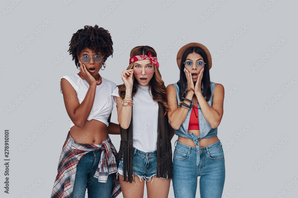 Poster three surprised young women making a face and looking at camera while standing against grey backgrou