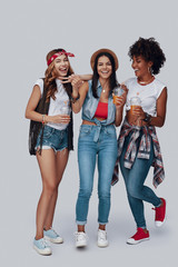 Full length of three attractive young women drinking refreshing cocktail and smiling while standing against grey background