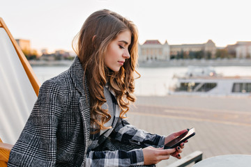 Pensive girl with curly hairstyle texting message, sitting on recliner in cold morning. Outdoor...