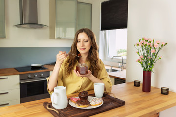 Young beautiful girl is having breakfast at home in the kitchen. She drinks her morning coffee and with great pleasure eats jam from a can.