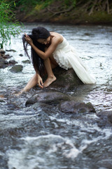 Young pretty brunette woman in white wedding dress sitting on the rock in rushing river