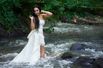 Young pretty brunette woman in white wedding dress stands outdoors in rushing river