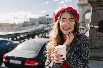 Curious blonde girl in elegant red beret posing with smile on blur background in windy morning....