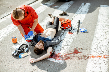 Ambluence worker applying emergency care with defibrillator to the injured bleeding man lying on the pedestrian crossing - obrazy, fototapety, plakaty