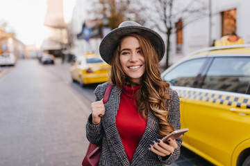 Pretty pale lady in casual attire hurrying to job in autumn morning. Smiling white woman in gray tweed jacket and hat walking down the street near yellow taxi and waiting for call.