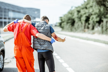 Medic in uniform helping injured man to walk, applying first aid after the road accident